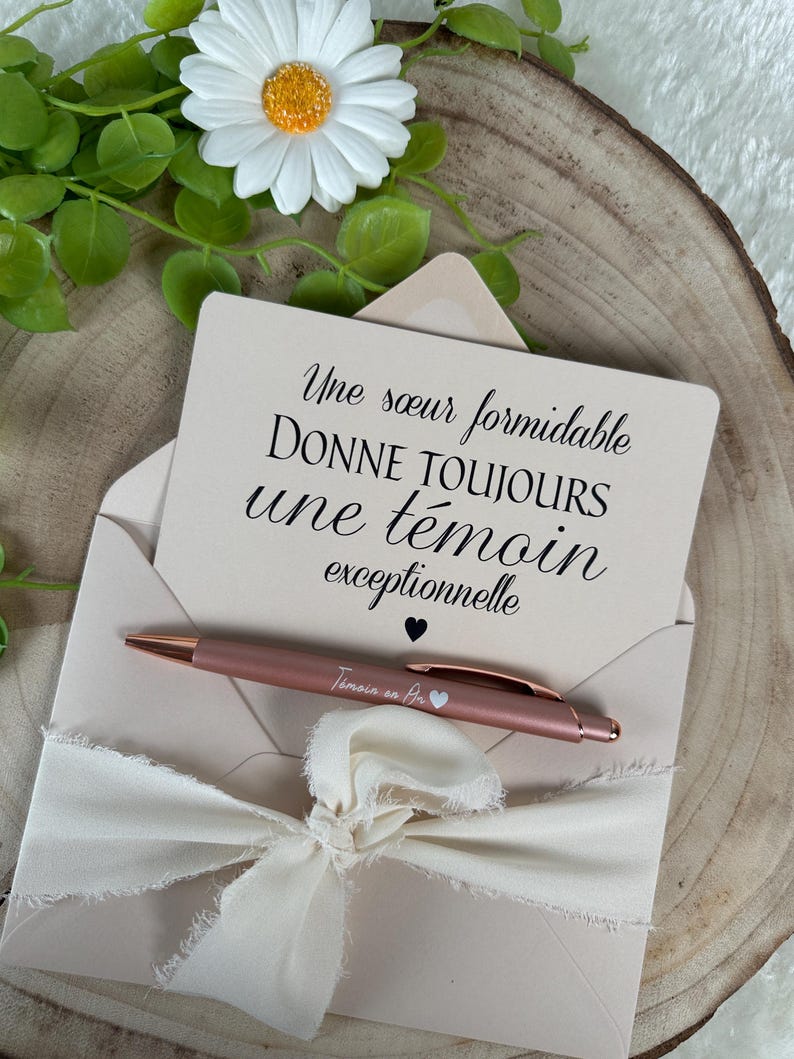 K&ouml;nnte beinhalten: Ein ros&eacute;goldener Stift und eine Karte mit franz&ouml;sischem Text werden in einem hellrosa Umschlag mit cremefarbenem Band pr&auml;sentiert. Auf der Karte steht: "Une s&oelig;ur formidable donne toujours une t&eacute;moin exceptionnelle."