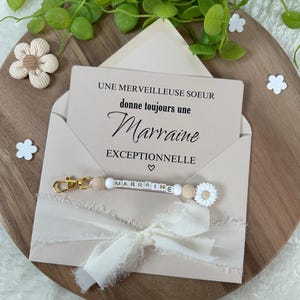 May include: A pink envelope with a white card inside that reads "Une merveilleuse soeur donne toujours une Marraine exceptionnelle". A white ribbon with a gold charm that says "Marraine" is attached to the envelope.
