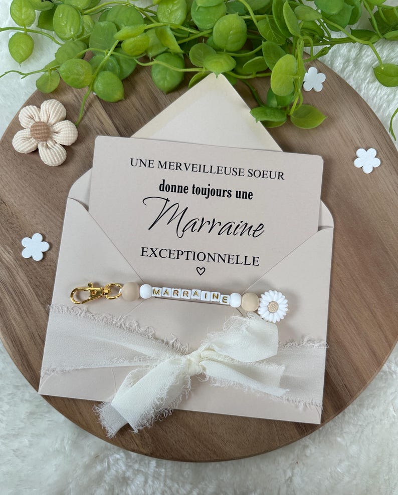 May include: A pink envelope with a white card inside that reads "Une merveilleuse soeur donne toujours une Marraine exceptionnelle". A white ribbon with a gold charm that says "Marraine" is attached to the envelope.