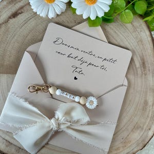 Puede incluir: Un sobre beige con una cinta color crema y un llavero que deletrea "TATA" con cuentas de madera y un dije de margarita. La tarjeta en el interior dice "Dans mon ventre, un petit coeur bat d&eacute;j&agrave; pour toi, Tata."