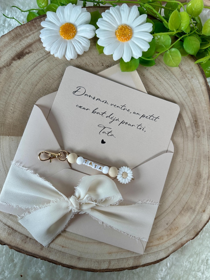 Puede incluir: Un sobre beige con una cinta color crema y un llavero que deletrea "TATA" con cuentas de madera y un dije de margarita. La tarjeta en el interior dice "Dans mon ventre, un petit coeur bat d&eacute;j&agrave; pour toi, Tata."