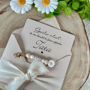 Puede incluir: Una tarjeta beige en un sobre con el texto "Spoiler alert: tu vas bient&ocirc;t te faire appeler. Tata". Un llavero con la palabra "TATA" est&aacute; adjunto. La tarjeta est&aacute; decorada con un lazo blanco y detalles de margaritas.