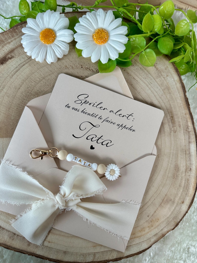 Puede incluir: Una tarjeta beige en un sobre con el texto "Spoiler alert: tu vas bient&ocirc;t te faire appeler. Tata". Un llavero con la palabra "TATA" est&aacute; adjunto. La tarjeta est&aacute; decorada con un lazo blanco y detalles de margaritas.