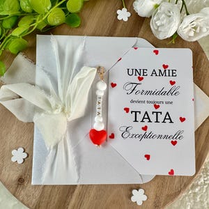 以下が含まれることがあります： 白いリボンで結ばれた白い封筒。カードには「UNE AMIE Formidable devient toujours une TATA Exceptionnelle」というテキストと赤いハートのアクセントが施されています。「TATA」という文字と赤いハートが付いたチャームが封筒からぶら下がっています。