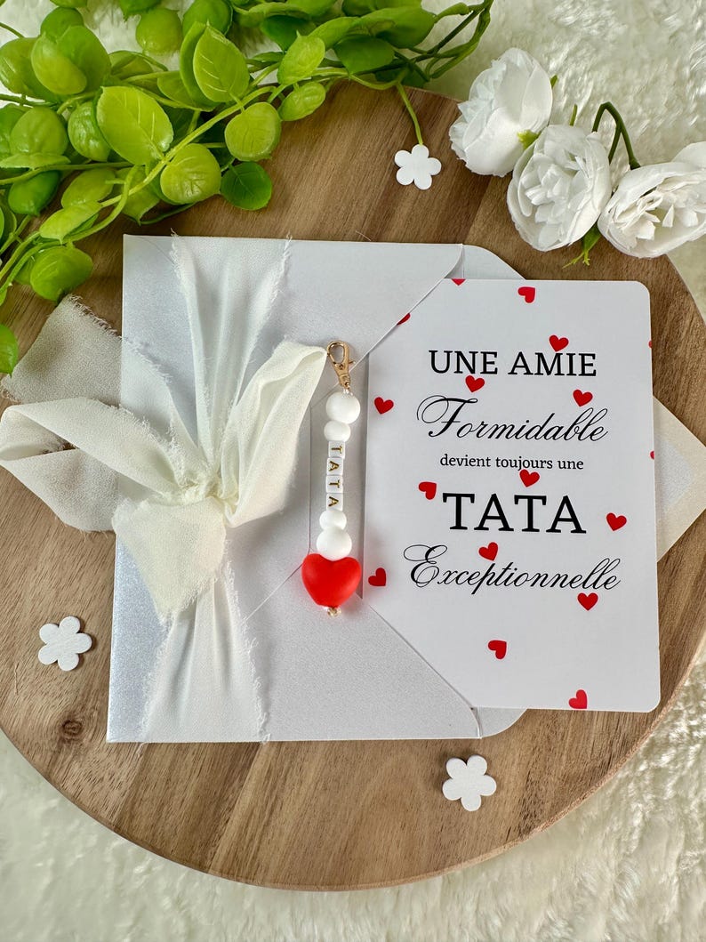 以下が含まれることがあります： 白いリボンで結ばれた白い封筒。カードには「UNE AMIE Formidable devient toujours une TATA Exceptionnelle」というテキストと赤いハートのアクセントが施されています。「TATA」という文字と赤いハートが付いたチャームが封筒からぶら下がっています。