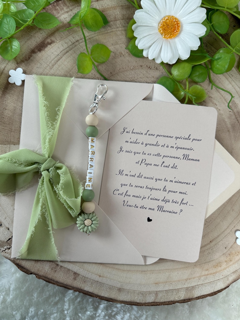May include: A white envelope with a green ribbon and a green and white beaded keychain with the word "MARRAINE" spelled out on it. The envelope has a handwritten note inside that reads "J'ai besoin d'une personne sp&eacute;ciale pour m'aider &agrave; grandir et &agrave; m'&eacute;panouir. Je sais que tu es cette personne, Maman et Papa me l'ont dit. Ils m'ont dit aussi que tu m'aimeras et que tu seras toujours l&agrave; pour moi. C'est fou mais je t'aime d&eacute;j&agrave; tr&egrave;s fort.... Veux-tu &ecirc;tre ma Marraine?"