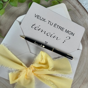 May include: An invitation card inside a white envelope, with a black pen and a yellow ribbon. The card reads "VEUX- TU &Ecirc;TRE MON t&eacute;moin ?" in black script. The pen is black with gold accents and the words "T&eacute;moins en Or" are written on the pen.