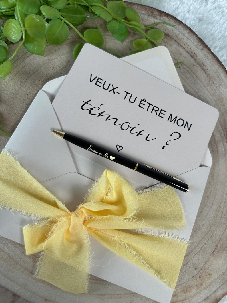 May include: An invitation card inside a white envelope, with a black pen and a yellow ribbon. The card reads "VEUX- TU &Ecirc;TRE MON t&eacute;moin ?" in black script. The pen is black with gold accents and the words "T&eacute;moins en Or" are written on the pen.