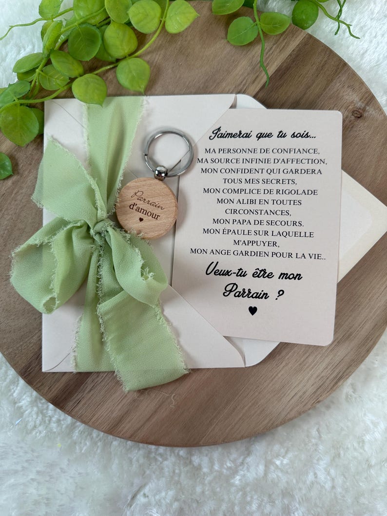 May include: A wooden keychain with the words "Parrain d'amour" engraved on it, attached to a light green ribbon and a card with French text. The card is inside a cream-colored envelope, with a wooden background.