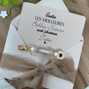 May include: A white envelope with a card inside that reads "Seules les meilleures Belles-Soeurs sont promues Tata". A brown ribbon with a beaded keychain that says "TATA" is attached to the envelope.