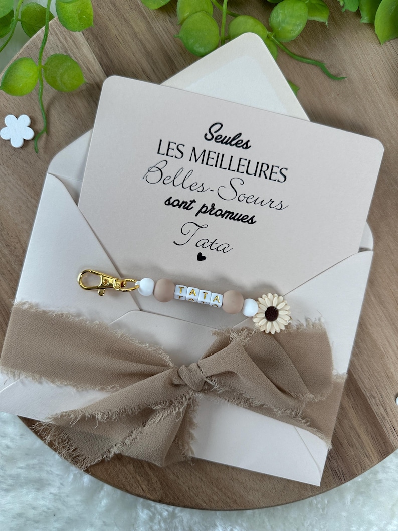 May include: A white envelope with a card inside that reads "Seules les meilleures Belles-Soeurs sont promues Tata". A brown ribbon with a beaded keychain that says "TATA" is attached to the envelope.