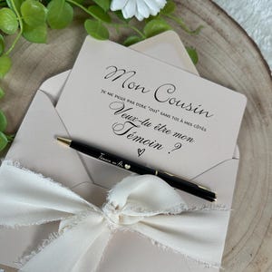 May include: A light pink invitation card with the text "Mon Cousin" and a request to be a witness, inside a matching envelope. A black pen with gold accents and the text "Temoin en Or" rests on the card, tied with a cream ribbon.