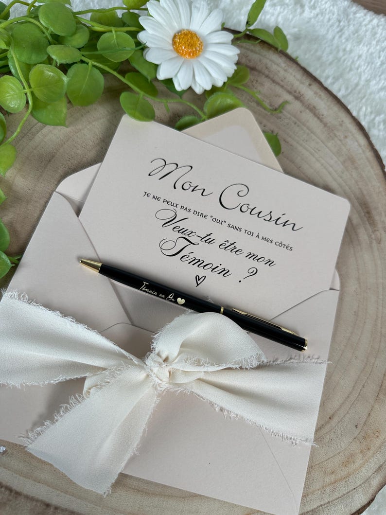 May include: A light pink invitation card with the text "Mon Cousin" and a request to be a witness, inside a matching envelope. A black pen with gold accents and the text "Temoin en Or" rests on the card, tied with a cream ribbon.
