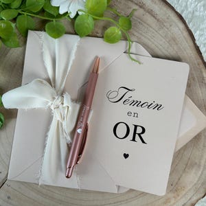 K&ouml;nnte beinhalten: Ein blushfarbenes Umschlag- und Kartenset, gebunden mit einem cremefarbenen Band. Ein ros&eacute;goldener Stift liegt auf dem Umschlag. Die Karte tr&auml;gt die Aufschrift "T&eacute;moin en OR" mit einem kleinen Herz. K&uuml;nstliche Blumen und eine Holzscheibe im Hintergrund.