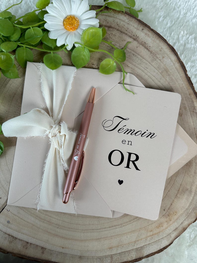 K&ouml;nnte beinhalten: Ein blushfarbenes Umschlag- und Kartenset, gebunden mit einem cremefarbenen Band. Ein ros&eacute;goldener Stift liegt auf dem Umschlag. Die Karte tr&auml;gt die Aufschrift "T&eacute;moin en OR" mit einem kleinen Herz. K&uuml;nstliche Blumen und eine Holzscheibe im Hintergrund.