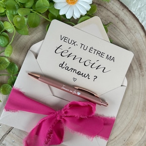 K&ouml;nnte beinhalten: Ein ros&eacute;goldener Stift und eine Karte in einem cremefarbenen Umschlag, gebunden mit einem leuchtend rosa Band. Auf der Karte steht "VEUX-TU &Ecirc;TRE MA t&eacute;moin d'amour ?" mit einem Herzsymbol. Eine wei&szlig;e G&auml;nsebl&uuml;mchen und Gr&uuml;nzeug im Hintergrund.