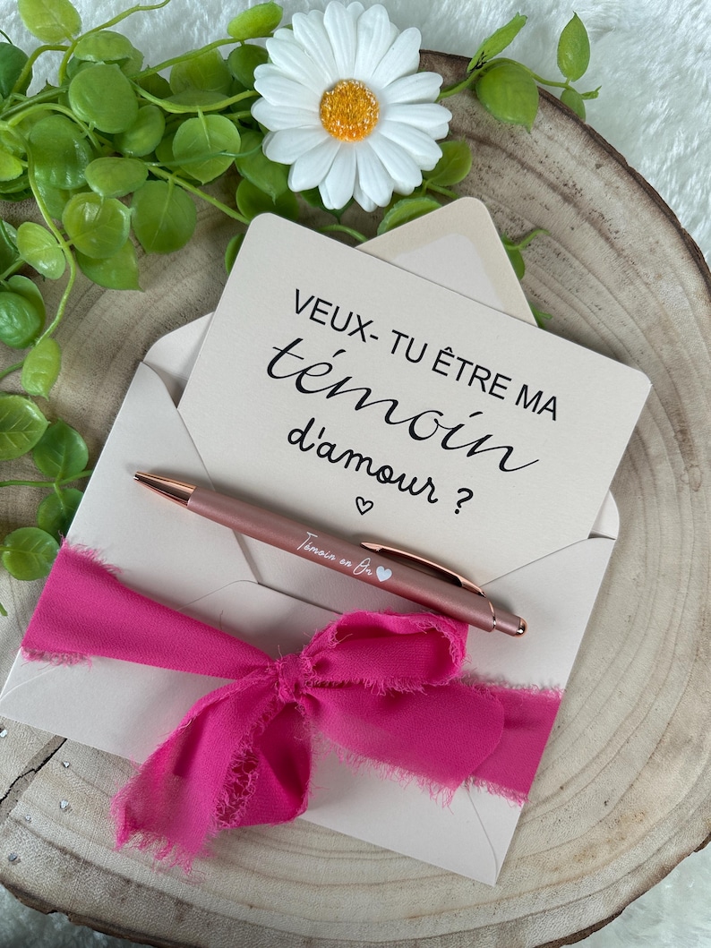 K&ouml;nnte beinhalten: Ein ros&eacute;goldener Stift und eine Karte in einem cremefarbenen Umschlag, gebunden mit einem leuchtend rosa Band. Auf der Karte steht "VEUX-TU &Ecirc;TRE MA t&eacute;moin d'amour ?" mit einem Herzsymbol. Eine wei&szlig;e G&auml;nsebl&uuml;mchen und Gr&uuml;nzeug im Hintergrund.