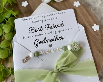 Puede incluir: Una tarjeta blanca con una cinta verde y una pulsera de cuentas con la palabra "Madrina" escrita. La tarjeta dice "Lo único que es mejor que tenerte como mejor amiga es que nuestro bebé te tenga como madrina."