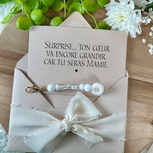 Pu&ograve; includere: Un biglietto rosa chiaro in una busta con il testo "SURPRISE... TON C&OElig;UR VA ENCORE GRANDIR, CAR TU SERAS MAMIE." Un portachiavi di perline bianche con la parola "MAMIE" e un ciondolo a forma di fiore bianco &egrave; attaccato. Un fiocco color crema adorna la busta.