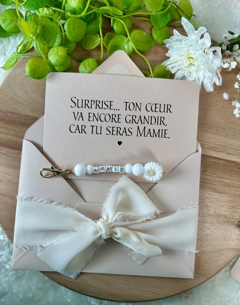 Pu&ograve; includere: Un biglietto rosa chiaro in una busta con il testo "SURPRISE... TON C&OElig;UR VA ENCORE GRANDIR, CAR TU SERAS MAMIE." Un portachiavi di perline bianche con la parola "MAMIE" e un ciondolo a forma di fiore bianco &egrave; attaccato. Un fiocco color crema adorna la busta.