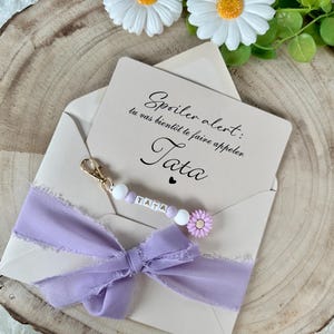 May include: A light beige envelope with a lavender ribbon and a beaded keychain that spells "TATA". The card inside reads "Spoiler alert: tu vas bient&ocirc;t te faire appeler Tata". Decorative daisies and greenery are in the background.