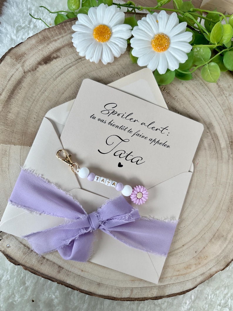 May include: A light beige envelope with a lavender ribbon and a beaded keychain that spells "TATA". The card inside reads "Spoiler alert: tu vas bient&ocirc;t te faire appeler Tata". Decorative daisies and greenery are in the background.