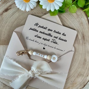 Puede incluir: Un sobre beige con un lazo crema contiene una tarjeta y un llavero. La tarjeta tiene texto en franc&eacute;s. El llavero tiene cuentas de madera con la palabra "TATA" y un dije de margarita blanca.