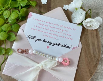 Puede incluir: Un sobre rosa con una cinta blanca y una tarjeta que dice "Necesito un par de manos extra para ayudarme a aprender y crecer. Sé que las tuyas serán las mejores porque mamá y papá me lo dijeron... ¿Quieres ser mi madrina?" Un llavero de cuentas rosa y blanco con la palabra "MADRINA" escrita en cuentas blancas.