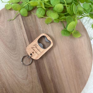May include: A wooden bottle opener with a silver metal opener and a keyring. The opener has the words "Parrain d'amour" engraved on it. The opener is on a wooden surface with green leaves in the background.
