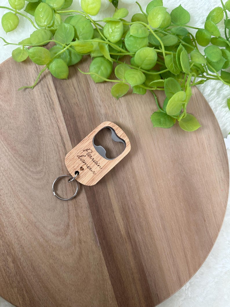 May include: A wooden bottle opener with a silver metal opener and a keyring. The opener has the words "Parrain d'amour" engraved on it. The opener is on a wooden surface with green leaves in the background.