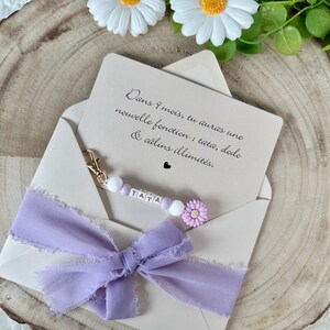 May include: A light beige envelope with a lavender ribbon and a beaded keychain that spells "TATA". The card inside reads "Dans 9 mois, tu auras une nouvelle fonction: tata, dodo & c&acirc;lins illimit&eacute;s." Decorative daisies are in the background.