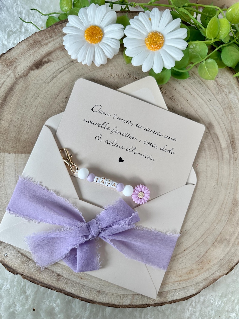 May include: A light beige envelope with a lavender ribbon and a beaded keychain that spells "TATA". The card inside reads "Dans 9 mois, tu auras une nouvelle fonction: tata, dodo & c&acirc;lins illimit&eacute;s." Decorative daisies are in the background.