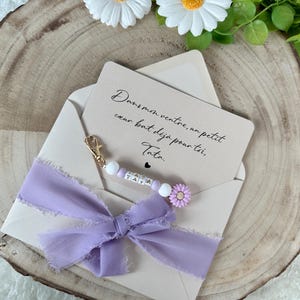 May include: A light beige envelope with a handwritten message and a lavender ribbon. The message reads, "Dans mon ventre, un petit coeur bat d&eacute;j&agrave; pour toi, Tata." A beaded charm with the word "TATA" is attached.