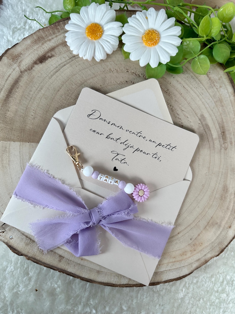May include: A light beige envelope with a handwritten message and a lavender ribbon. The message reads, "Dans mon ventre, un petit coeur bat d&eacute;j&agrave; pour toi, Tata." A beaded charm with the word "TATA" is attached.