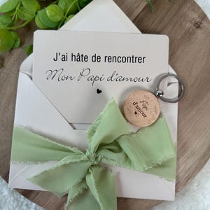 May include: A light pink envelope with a green ribbon bow, holding a card with French text. A wooden keychain with the words "Papi d'amour" is attached. The card reads "J'ai h&acirc;te de rencontrer Mon Papi d'amour."