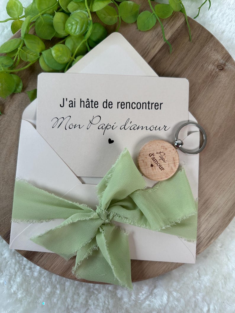 May include: A light pink envelope with a green ribbon bow, holding a card with French text. A wooden keychain with the words "Papi d'amour" is attached. The card reads "J'ai h&acirc;te de rencontrer Mon Papi d'amour."