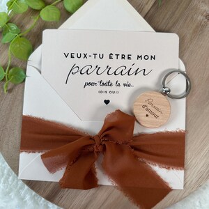 May include: A white card inside a white envelope with a brown ribbon tied around it. The card reads "VEUX-TU &Ecirc;TRE MON parrain pour toute la vie... (DIS OUI)". A wooden keychain with the words "Parrain d'amour" is also included.
