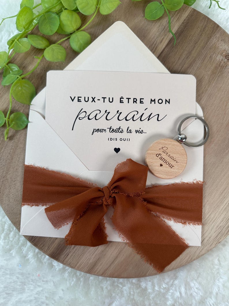May include: A white card inside a white envelope with a brown ribbon tied around it. The card reads "VEUX-TU &Ecirc;TRE MON parrain pour toute la vie... (DIS OUI)". A wooden keychain with the words "Parrain d'amour" is also included.