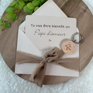 May include: A cream-colored envelope with a beige ribbon and a card that reads "Tu vas &ecirc;tre bient&ocirc;t un Papi d'amour" is displayed. A wooden keychain with the words "Papi d'amour" is attached. The items are arranged on a wooden tray.