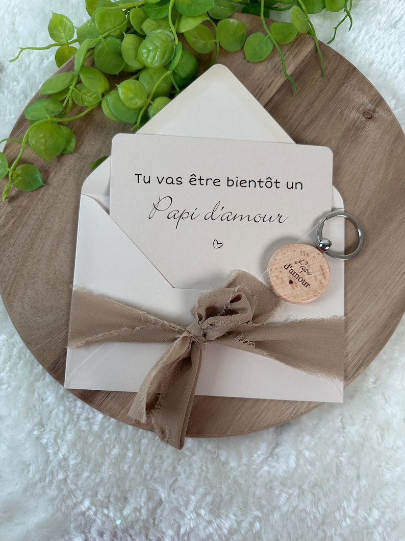 May include: A cream-colored envelope with a beige ribbon and a card that reads "Tu vas &ecirc;tre bient&ocirc;t un Papi d'amour" is displayed. A wooden keychain with the words "Papi d'amour" is attached. The items are arranged on a wooden tray.