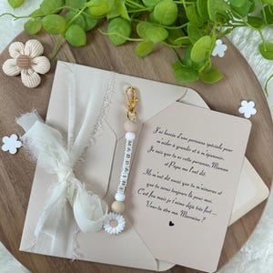 May include: A white card with a handwritten message in French asking "Veux-tu &ecirc;tre ma Marraine?" (Do you want to be my godmother?) with a white beaded keychain with the word "Marraine" spelled out in gold letters.
