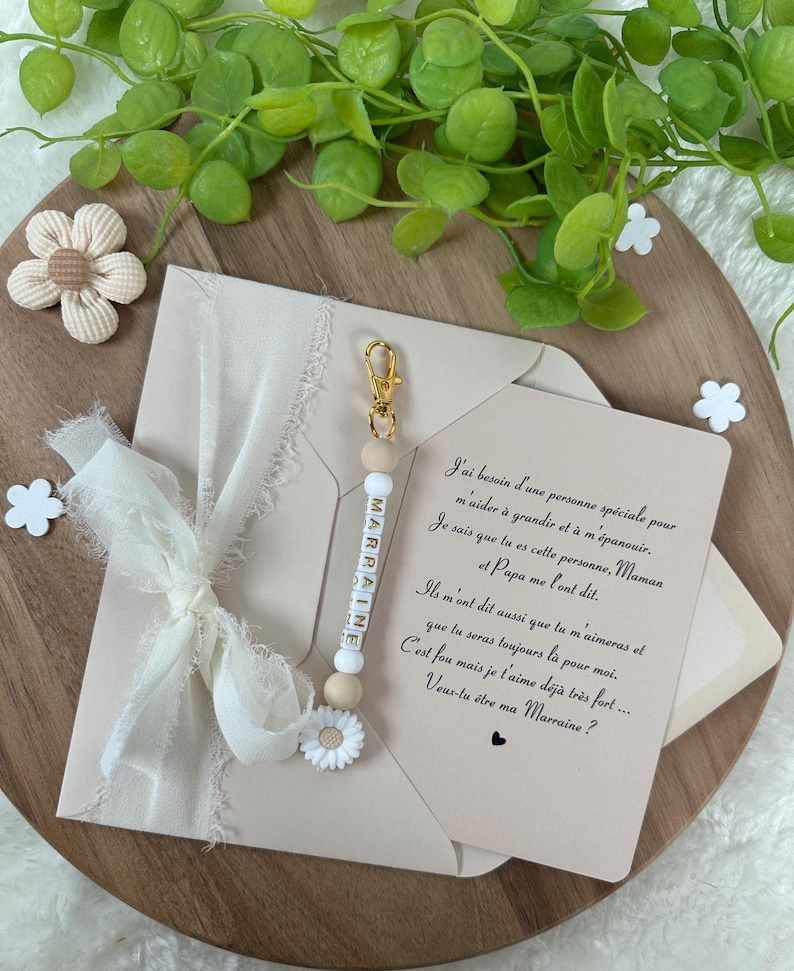 May include: A white card with a handwritten message in French asking "Veux-tu &ecirc;tre ma Marraine?" (Do you want to be my godmother?) with a white beaded keychain with the word "Marraine" spelled out in gold letters.