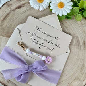 May include: A light purple ribbon bow and a keychain with the word "TATA" on it, attached to a beige envelope. The keychain has white beads and a small purple flower. The envelope contains a card with handwritten text.