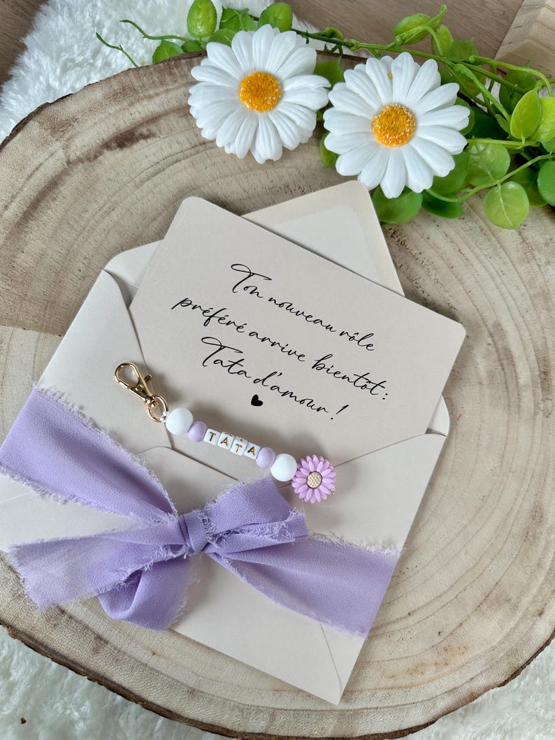 May include: A light purple ribbon bow and a keychain with the word "TATA" on it, attached to a beige envelope. The keychain has white beads and a small purple flower. The envelope contains a card with handwritten text.