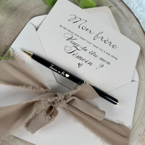 May include: A light pink envelope with a card inside, featuring elegant script that reads "Mon fr&egrave;re, Veux-tu &ecirc;tre mon T&eacute;moin?" A black pen with gold accents and the text "T&eacute;moin en Bo" rests on the envelope, tied with a beige ribbon.