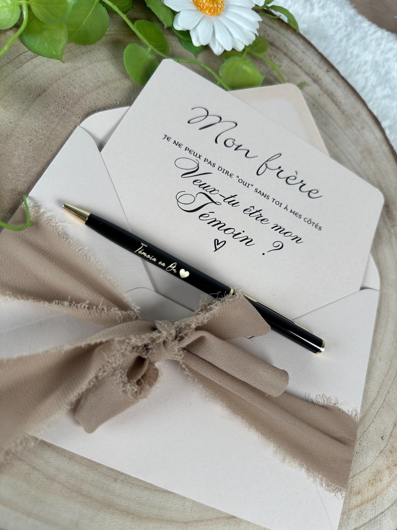 May include: A light pink envelope with a card inside, featuring elegant script that reads "Mon fr&egrave;re, Veux-tu &ecirc;tre mon T&eacute;moin?" A black pen with gold accents and the text "T&eacute;moin en Bo" rests on the envelope, tied with a beige ribbon.