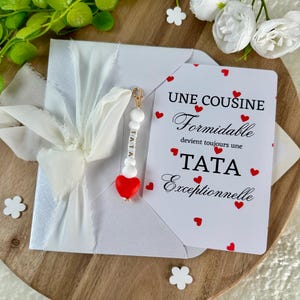 以下が含まれることがあります： 白いリボンで結ばれた白い封筒。カードには「UNE COUSINE Formidable devient toujours une TATA Exceptionnelle」というテキストと赤いハートが書かれています。「TATA」という文字と赤いハートが付いたチャームが付いています。