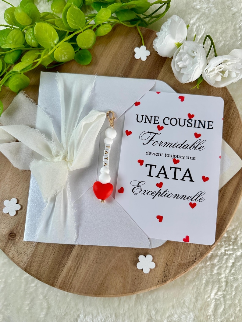 以下が含まれることがあります： 白いリボンで結ばれた白い封筒。カードには「UNE COUSINE Formidable devient toujours une TATA Exceptionnelle」というテキストと赤いハートが書かれています。「TATA」という文字と赤いハートが付いたチャームが付いています。