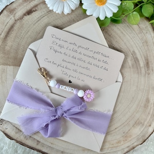 May include: A light beige envelope with a lilac ribbon and a beaded charm that says "TATA". The envelope contains a card with French text. Two white daisy flowers with yellow centers are at the top.