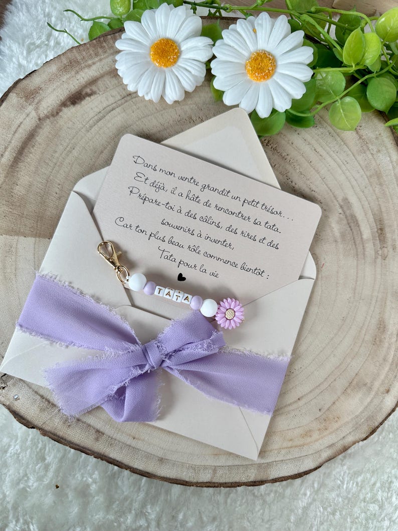 May include: A light beige envelope with a lilac ribbon and a beaded charm that says "TATA". The envelope contains a card with French text. Two white daisy flowers with yellow centers are at the top.